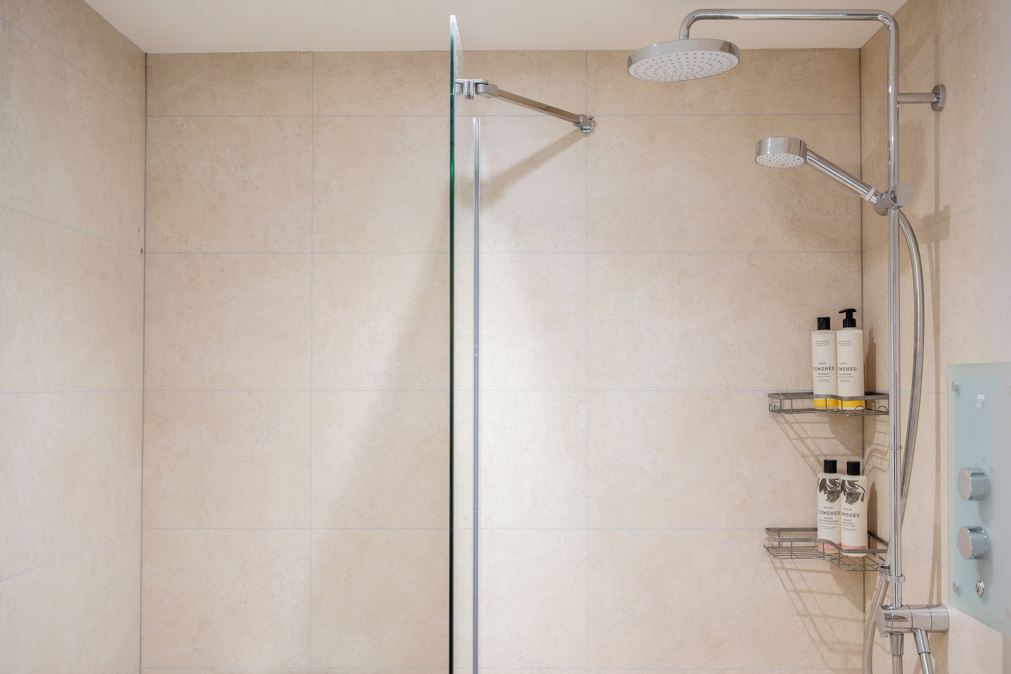 Een douche met een handdouche en een wandmengkraan, een glazen scheiding en planken met toiletartikelen.