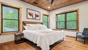 A bedroom with a wooden ceiling, large windows, a bed, a chair, and a framed picture on the wall.