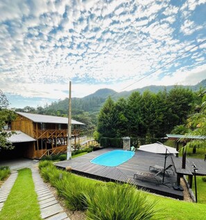 A pool area with a wooden deck, a covered seating area, and a view of a mountain range. - Sitío Rústico do Zizo com Piscina (Angelina)