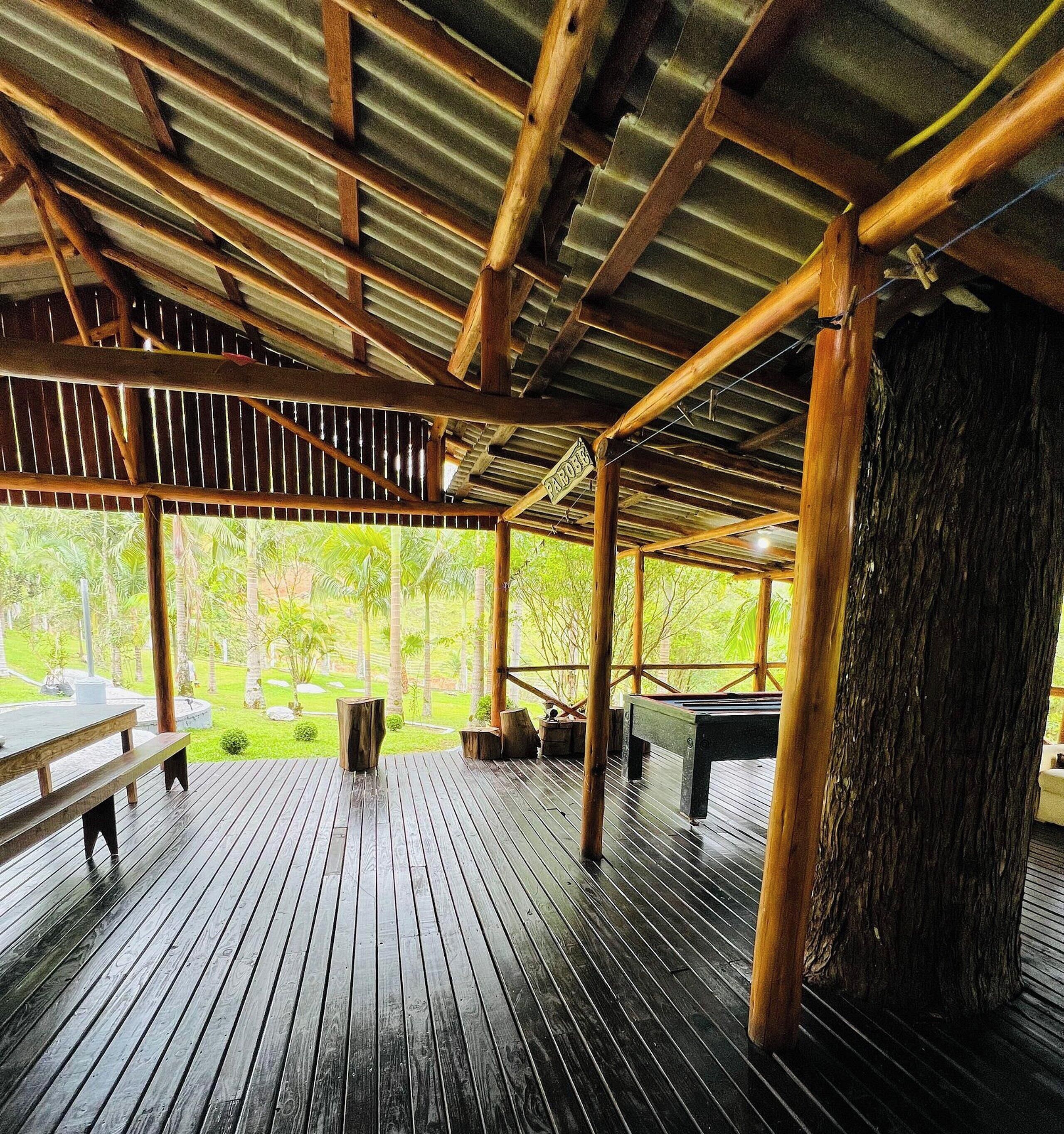 A covered wooden deck with a pool table, benches, and a view of a lush garden.