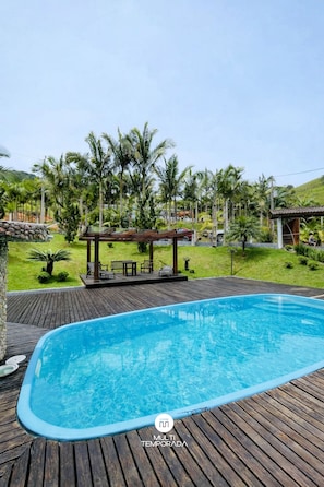 A swimming pool with a wooden deck, surrounded by greenery and a gazebo. - Sitío Rústico do Zizo com Piscina (Angelina)
