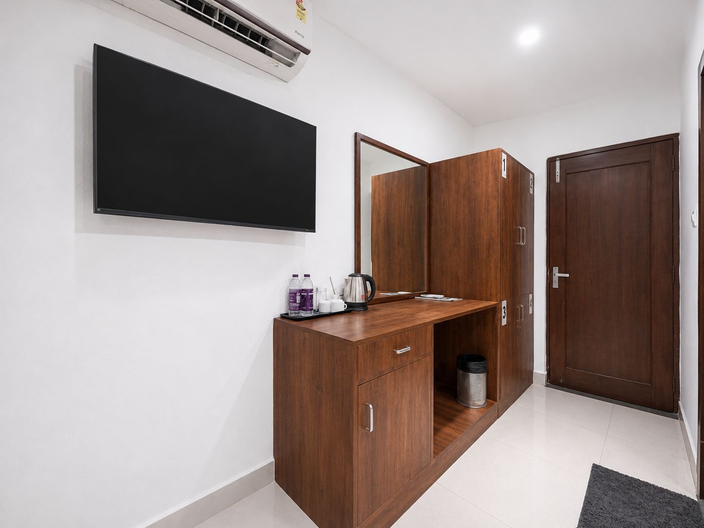 A hotel room with a flat-screen TV mounted on the wall, a wooden desk with a tray of toiletries, a mirror, and a wardrobe with a door marked '1'.