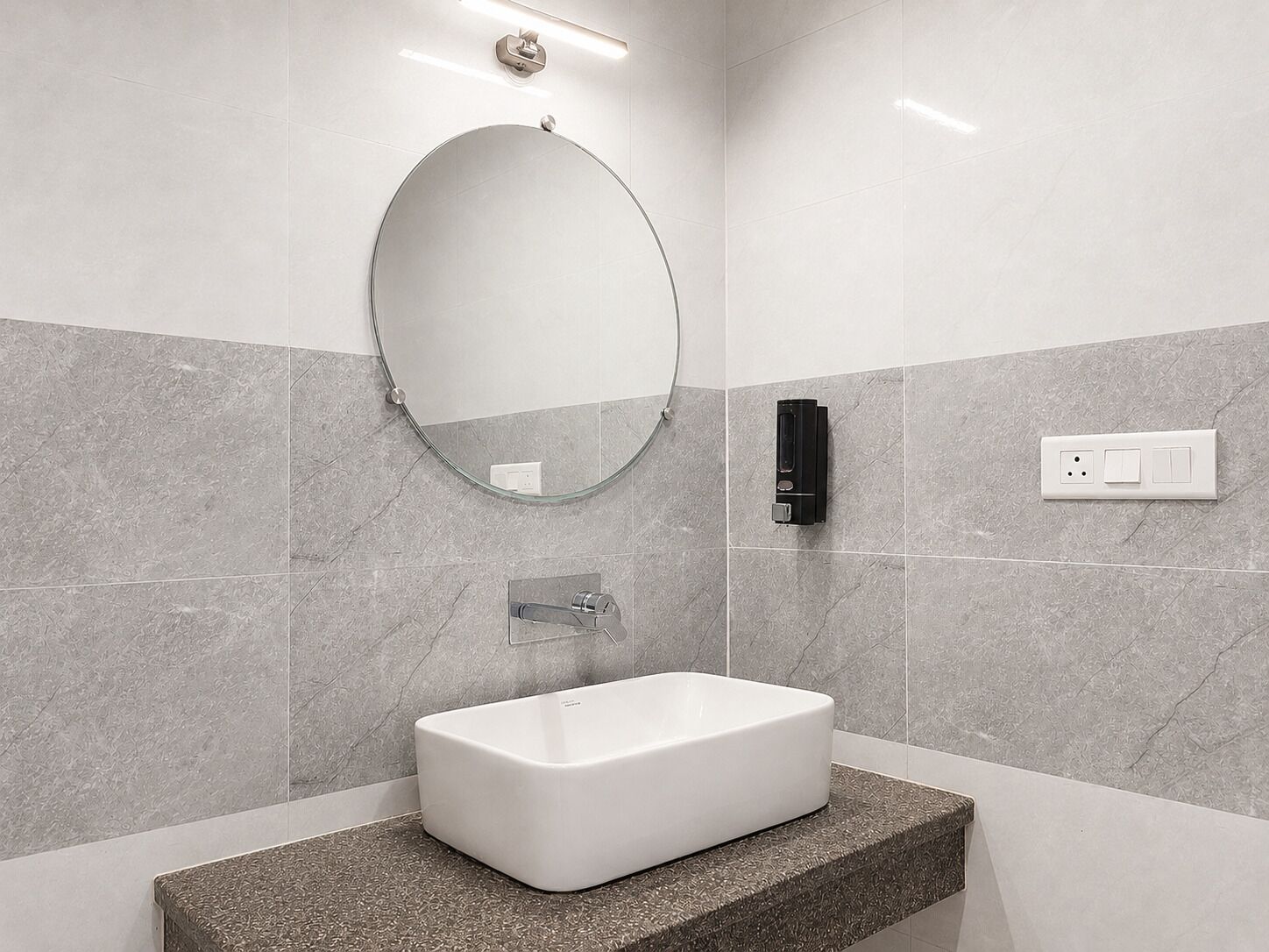 A bathroom with a rectangular sink, a round mirror, and a wall-mounted soap dispenser.