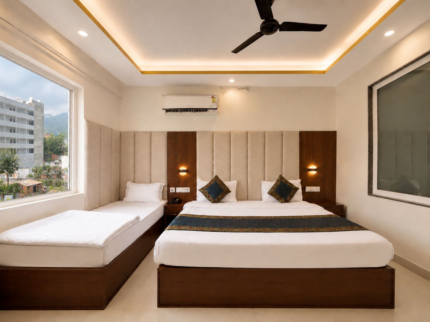 A hotel room with a bed, a smaller bed, a ceiling fan, and a window with a view of buildings.