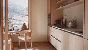 A modern kitchen with wooden cabinets, a dining area with a table and chairs, and a view of a snowy landscape through a large window. - DAS AICH - Suite Luma (Aich-Assach)
