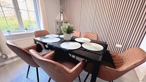 Spiseplads med et bord dækket til fire personer, omgivet af polstrede stole, en stueplante og en stribet væg.