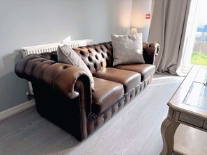 A brown leather sofa with tufted detailing, a white radiator, and a view of greenery outside. - Elegant 4BDR Home with Private Parking in Darfield (Darfield)