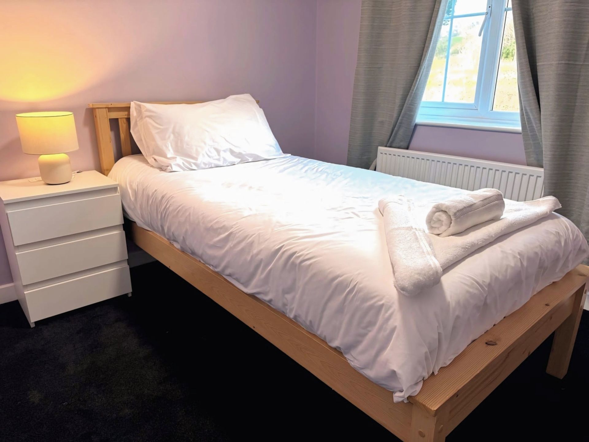 A neatly made bed with white linens, a bedside lamp, and a window with curtains.