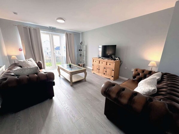 Una sala de estar con sofá, mesa de centro, mueble para televisión y balcón con cortinas.