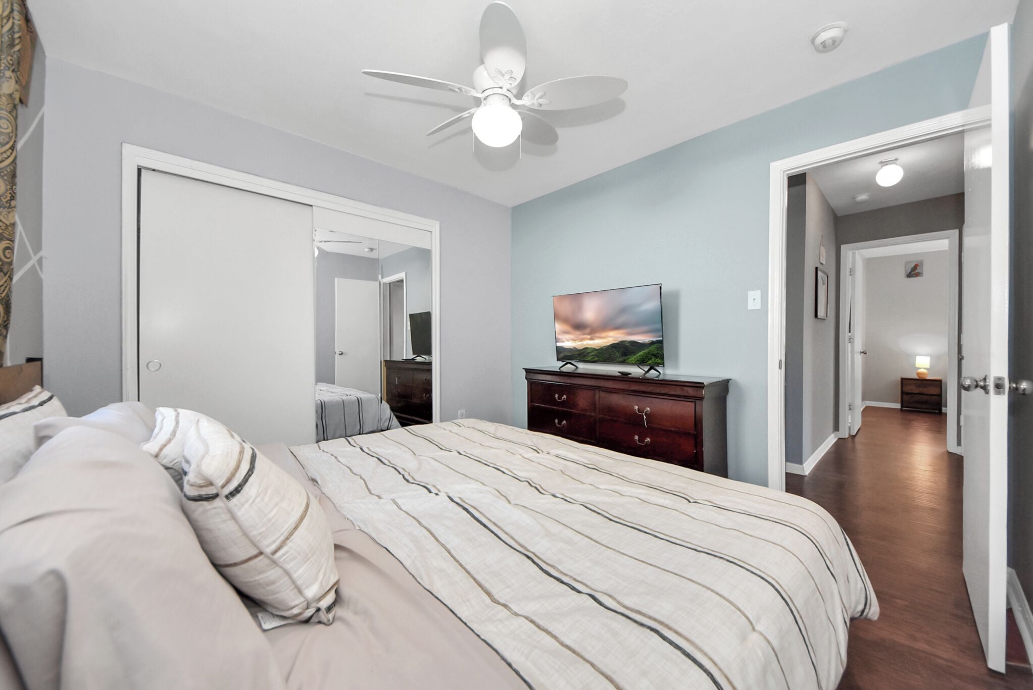 A bedroom with a bed, a ceiling fan, a TV, a dresser, and a hallway leading to another room.