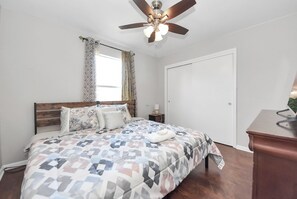Ein Schlafzimmer mit Bett, Deckenventilator, Fenster mit Vorhängen, einer Tür und einem kleinen Nachttisch.