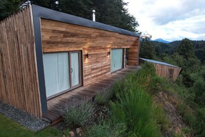 A modern wooden cabin with large windows, a deck, and a view of the surrounding forest.