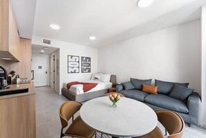 Et moderne hotelværelse med tekøkken, seng, grå sofa, et rundt spisebord med stole og et billedværk på væggen.