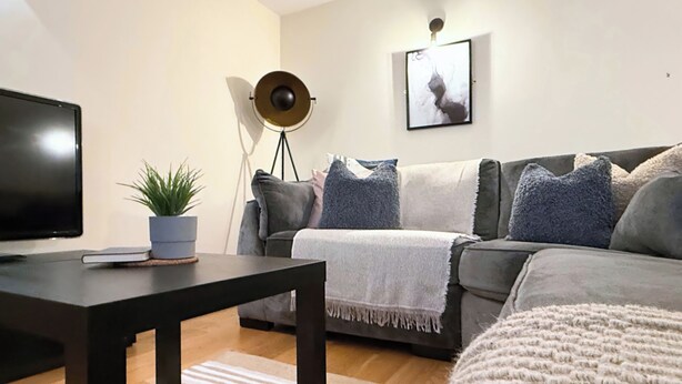 Una sala de estar con un televisor de pantalla plana, una mesa de centro oscura, una planta en maceta, un sofá gris con una manta y una lámpara de pie, además de un cuadro en la pared.
