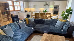 A living room with a sectional sofa, a wooden coffee table, and a kitchen in the background. - Old Corner Barn (Penrith)