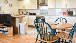 A modern kitchen with white cabinets, a stainless steel range hood, and a dining area with a wooden table and blue chairs. - Crag Barn Cottage (Penrith)