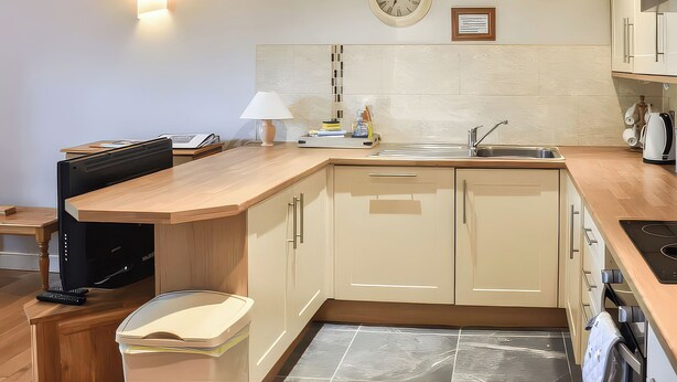 A modern kitchen with a wooden countertop, a sink, a clock, and a small table with a lamp. - Crag Barn Cottage (Penrith)
