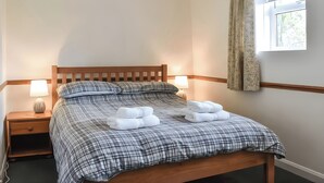 A neatly made bed with a checkered bedspread, two bedside lamps, a window with a curtain, and a wooden headboard. - Crag Barn Cottage (Penrith)