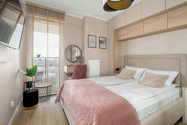 A sun-drenched bedroom features a large bed with a dusty rose throw and a view leading out to a private balcony. Built-in wooden cabinetry above the headboard maximizes storage in the compact, modern space.
