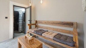 Un banc en bois avec des coussins, une table en bois avec des bols, et un espace douche visible à travers une porte ouverte.