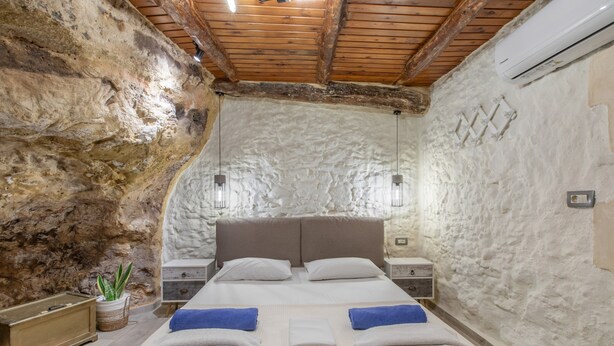 Een slaapkamer met een stenen muur, houten balken in het plafond, een bed met twee kussens, een nachtkastje en een wandairconditioner.