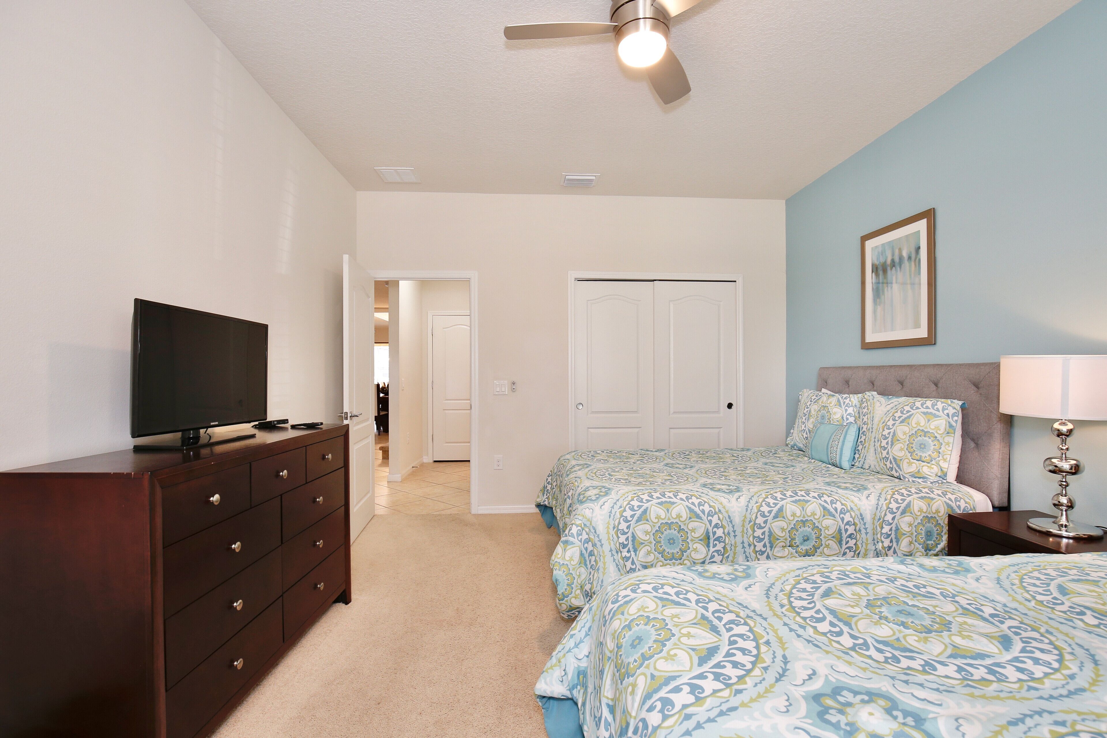 A bedroom with a TV on a dresser, a bed with patterned bedding, a ceiling fan, and a door leading to another room.