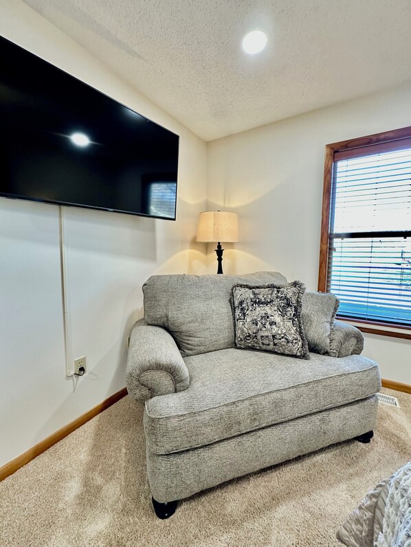 A living room with a grey sofa, a mounted TV, a lamp, and a window with blinds. - Private river access, at the gateway to the smokies in the heart of Townsend! (townsend)