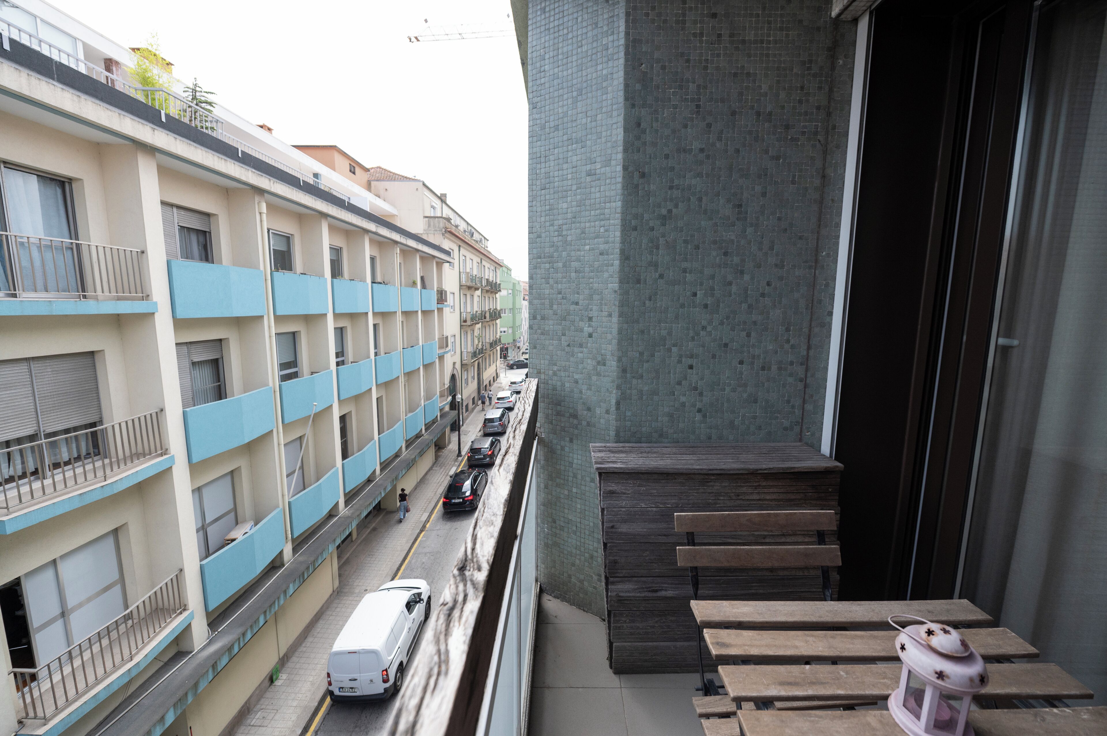 A multi-story building with balconies, a street with parked cars, and a wooden bench area.