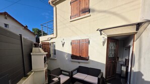 Eine Terrasse mit Gartenmöbeln, einer kleinen Küchenzeile und einer weißen Wand mit Holzjalousien.