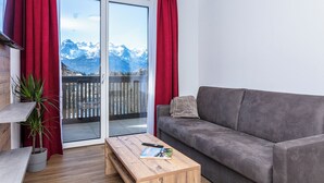 A living room with a grey sofa, a wooden coffee table with a remote control and a magazine, a potted plant, and a sliding glass door leading to a balcony with a mountain view. - Centrally located apartment hotel, right next to the cable car, wellness breakfast (Werfenweng)