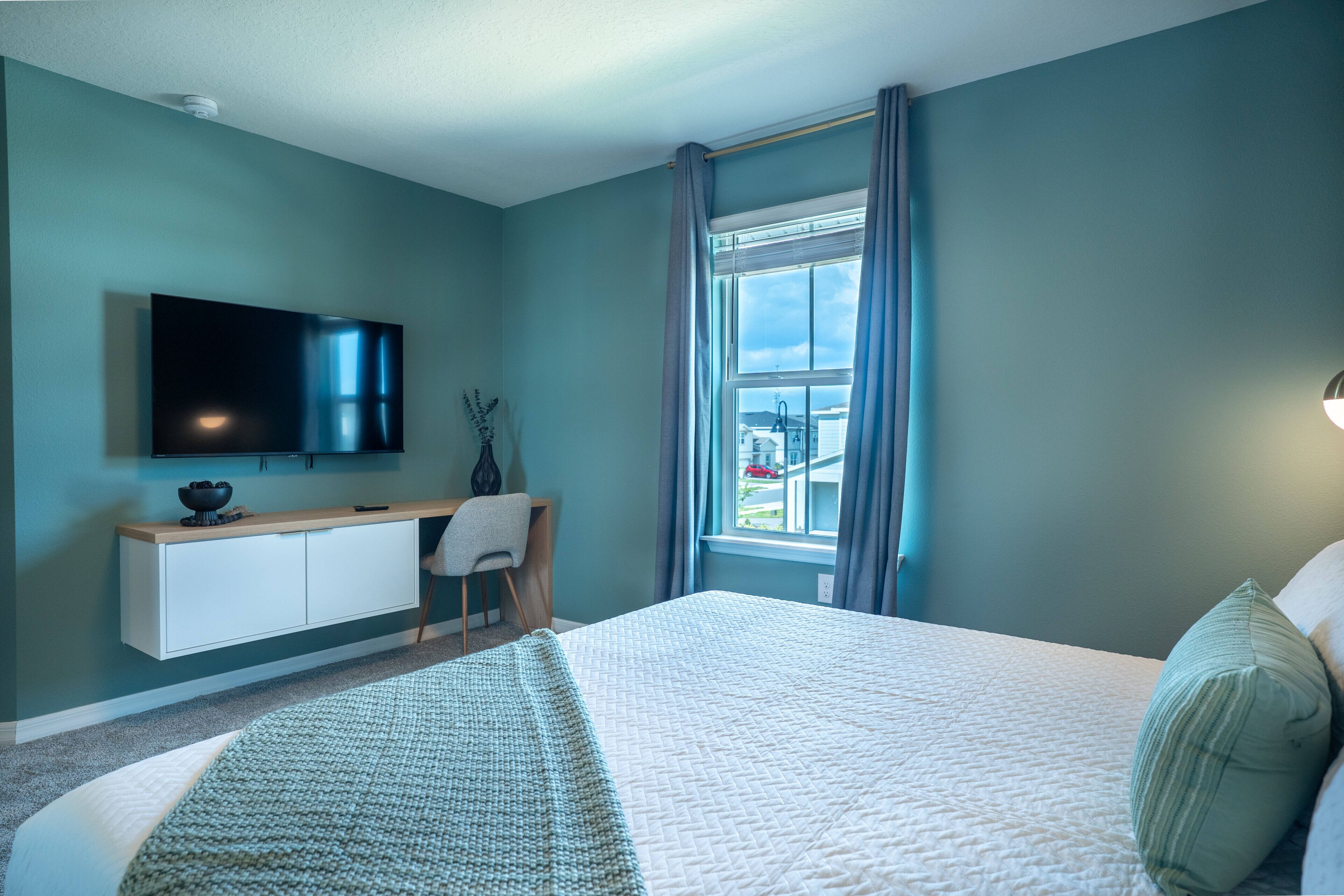 A bedroom with a bed, a TV mounted on the wall, a window with curtains, and a bedside table.
