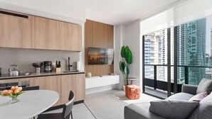 Une cuisine moderne avec des meubles en bois, un coin repas avec une table ronde et des chaises, une grande fenêtre donnant sur la ville et un canapé gris dans l’espace de vie.