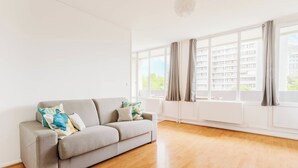 Amplia sala de estar con un sofá gris, piso de madera y grandes ventanas con cortinas.