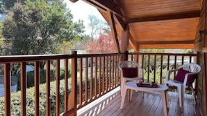 A wooden deck with two white plastic chairs and a small table, overlooking a garden with trees and a white fence.