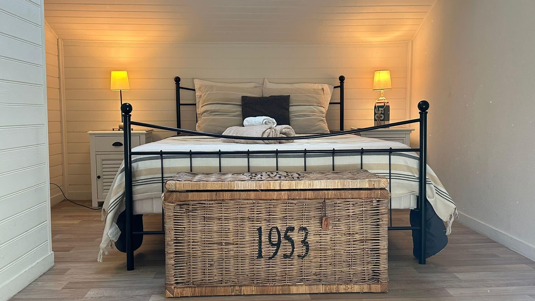 A bed with a metal frame, two bedside lamps, a bedside table, and a wicker basket with the number '1953' on the floor.
