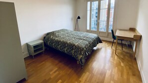 Una camera da letto con un letto, una scrivania, una sedia, una lampada da terra e una finestra vista edifici.
