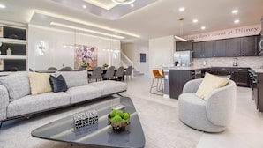 A modern living room with a sofa, armchair, and coffee table. There is a kitchen area with dark cabinets and a countertop. - Last Minute Disney Getaway! Private Pool, Game Room & Themed Bedrooms. Book Now! #163613 (Orlando)