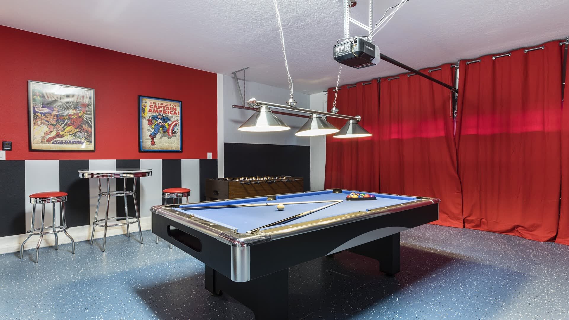 Een pooltafel met verlichting erboven, een rood gordijn en twee posters aan de muur.