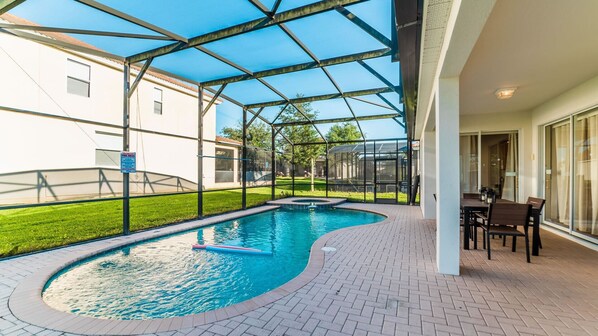 Une piscine entourée de parois en verre, un espace patio avec une table et des chaises, et une vue sur une maison en arrière-plan.