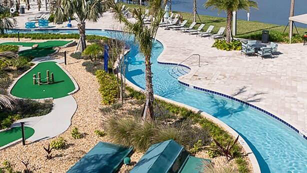 Un área de piscina con borde curvo, sillas de descanso, palmeras y vista a un cuerpo de agua.