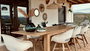 Ein Essbereich mit einem Holztisch und weißen Stühlen, umgeben von Steinböden, unter einer Holzdielendecke mit Pendelleuchten.