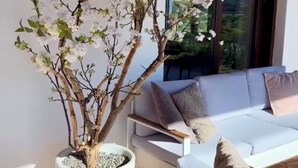 Ein modernes Wohnzimmer mit einem weißen Sofa, einer großen Topfpflanze und einem Fenster mit Blick auf Grünflächen.