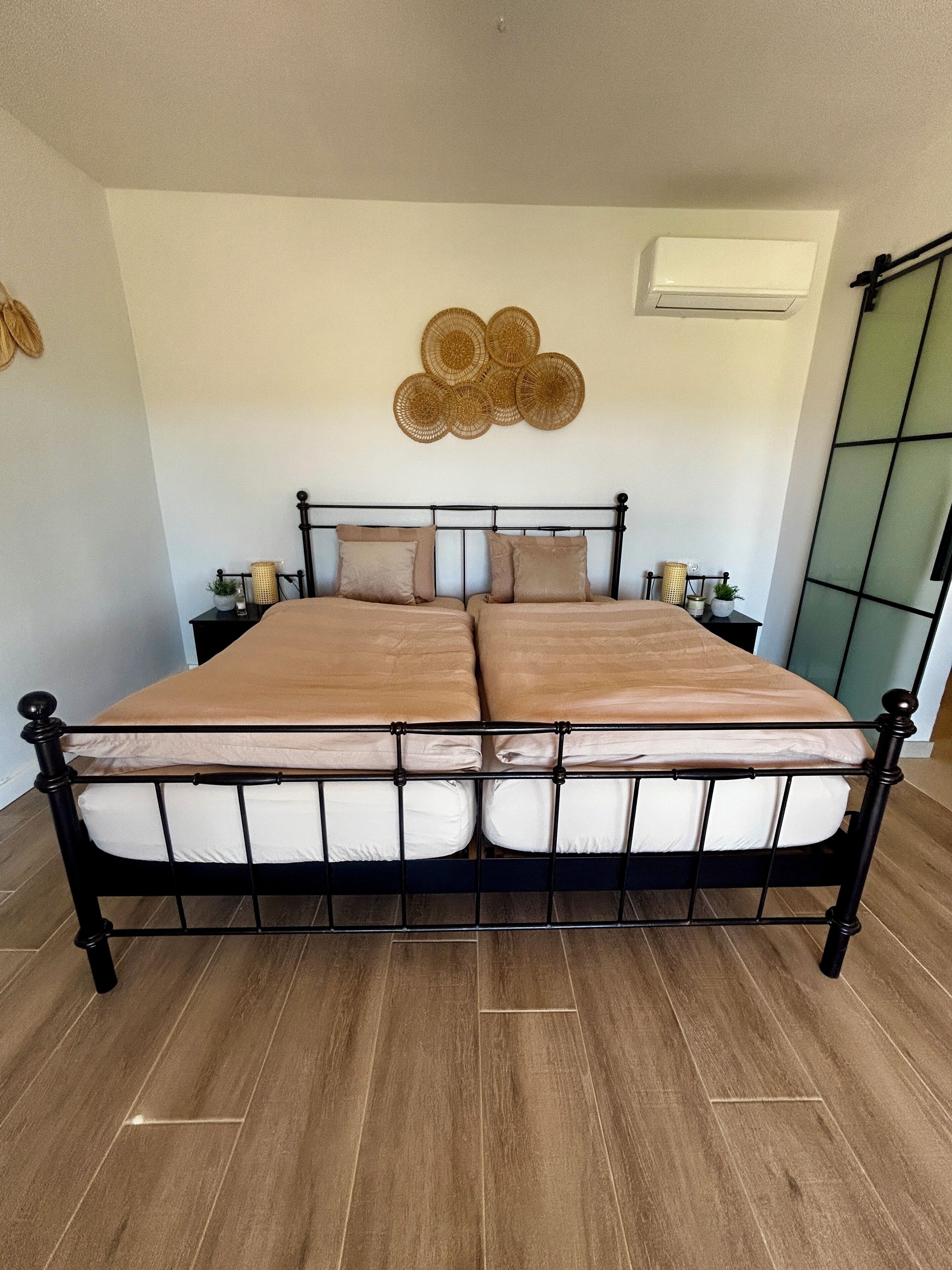 A double bed with a metal frame, two bedside tables with lamps, a wall-mounted air conditioner, and a decorative wall piece.