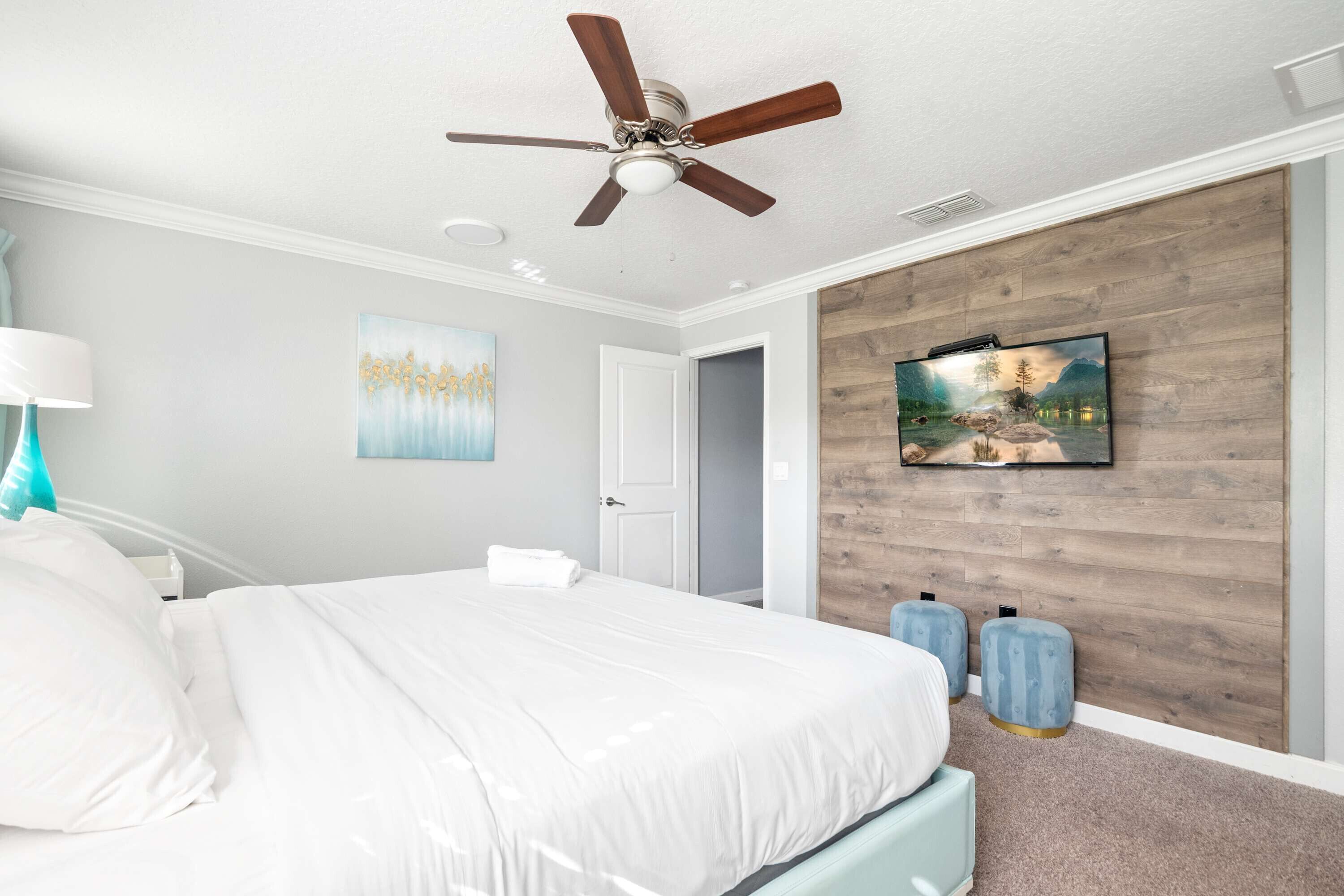 Une chambre à coucher avec un lit, un ventilateur de plafond, un téléviseur fixé au mur en bois, et une porte donnant sur une autre pièce.