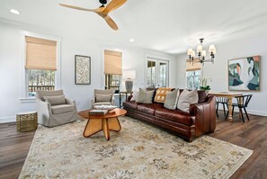 Uma sala de estar com um sofá de couro marrom, poltronas bege, uma mesa de centro e um espaço de jantar com mesa e cadeiras.