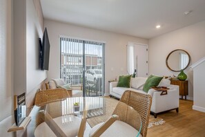 Ein modernes Wohnzimmer mit einem Glastisch, Korbstühlen, einem weißen Sofa mit grünen Kissen, einem großen Fenster mit Jalousien und einem runden Spiegel an der Wand.