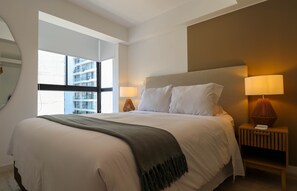 Una habitación de hotel moderna con una cama grande, dos lámparas de noche, una mesita de noche y vistas a un edificio a través de un ventanal amplio.