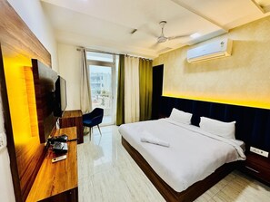 Quarto de hotel com cama, escrivaninha com televisão, cadeira, ventoinha no teto e ar condicionado.