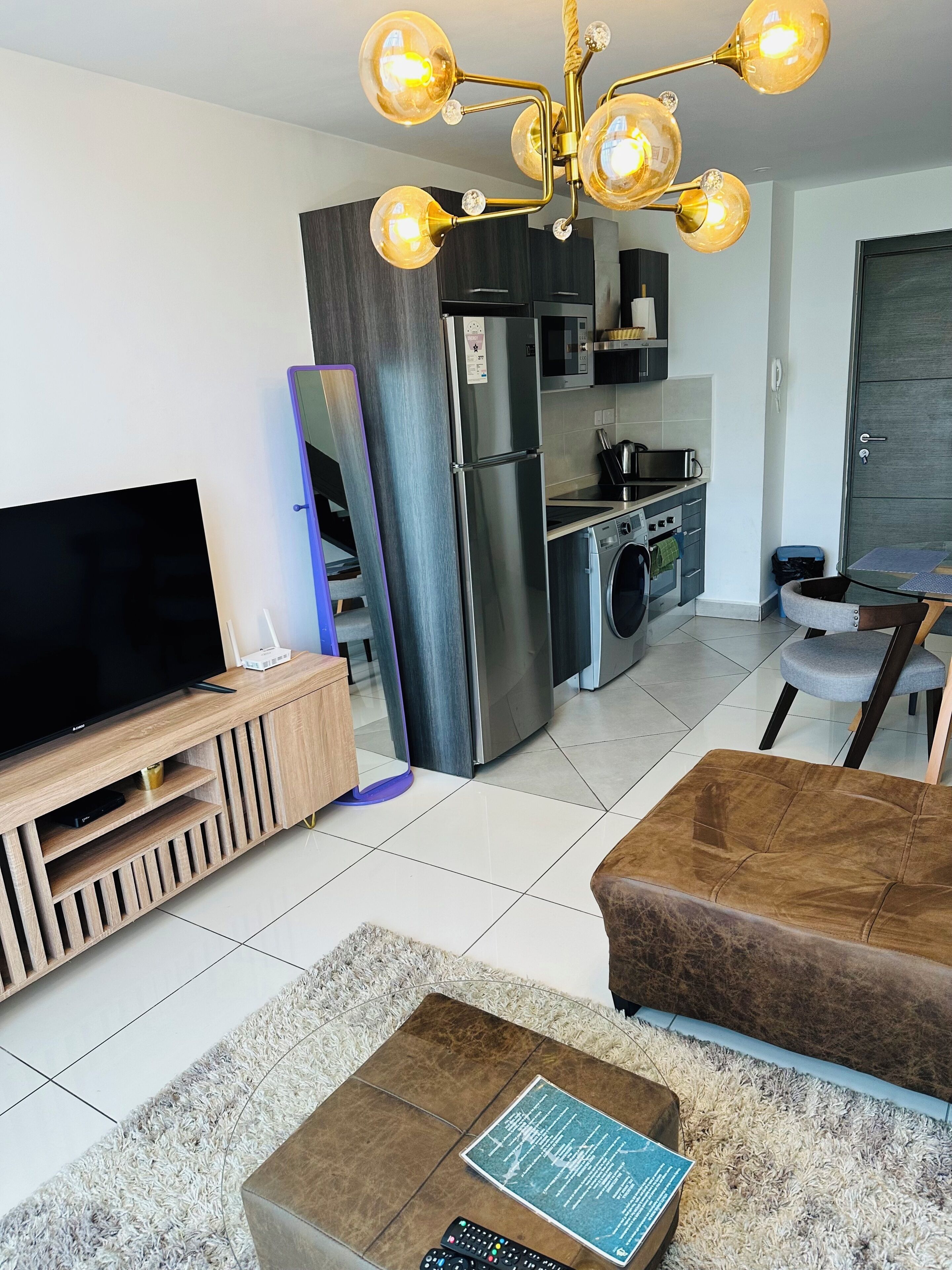 A modern living room with a flat-screen TV, a sofa, a coffee table with a book, a kitchen area with a refrigerator, microwave, and stove, and a dining area with a chair.