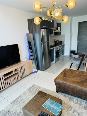 A modern living room with a flat-screen TV, a sofa, a coffee table with a book, a kitchen area with a refrigerator, microwave, and stove, and a dining area with a chair. - danny's lux home  (Accra)